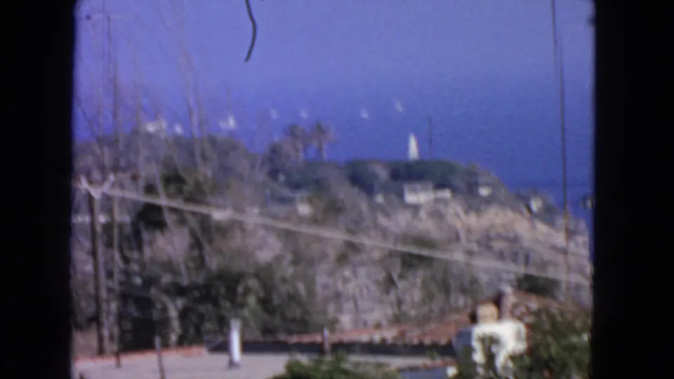 1963 8mm San Francisco coastal frame with brushy hillside, utility lines, rooftops, and a distant white tower against blue sky.