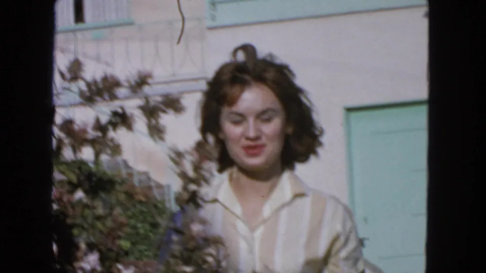 1963 San Francisco home-movie frame with a young woman in a light collared shirt smiling near shrubs and a mint-green door.