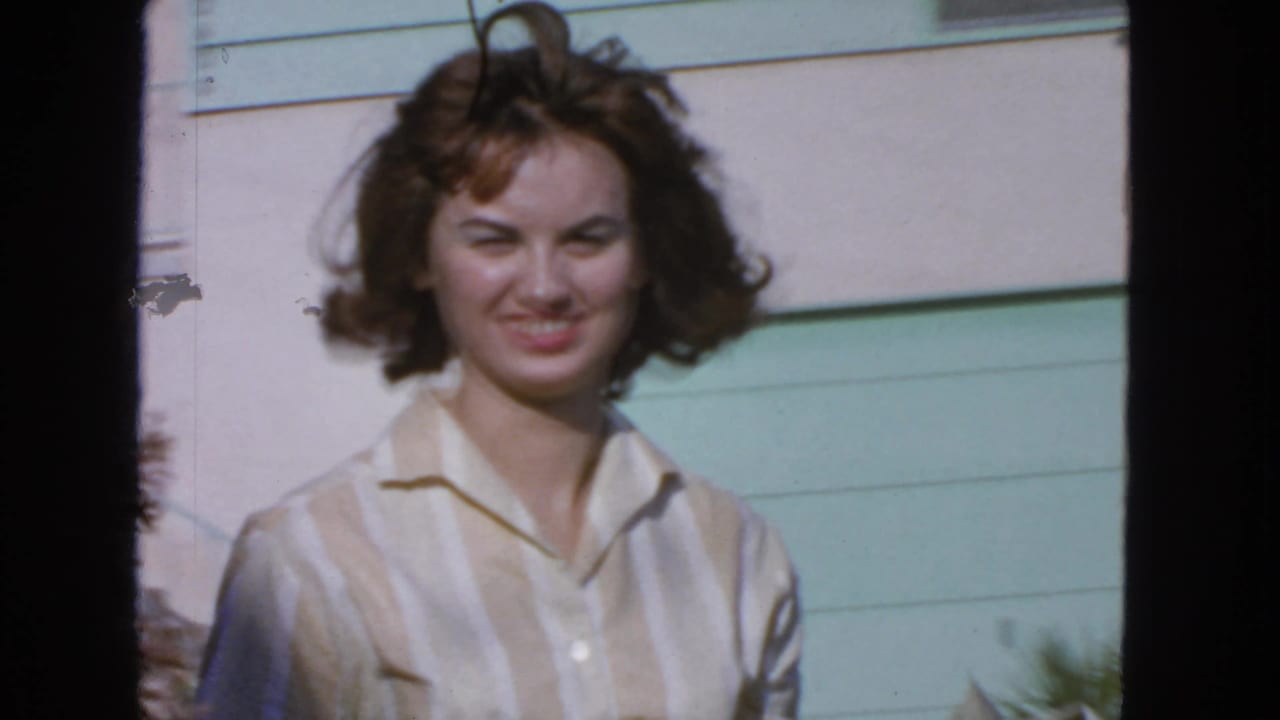 1963 San Francisco 8mm frame of a young woman smiling outdoors near pale siding, a mint-green surface, and plants.