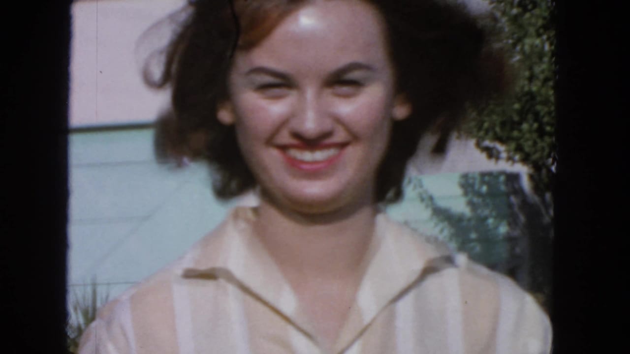 Closer 1963 San Francisco home-movie view of the smiling woman, with brown hair lifted by wind and garden foliage behind her.
