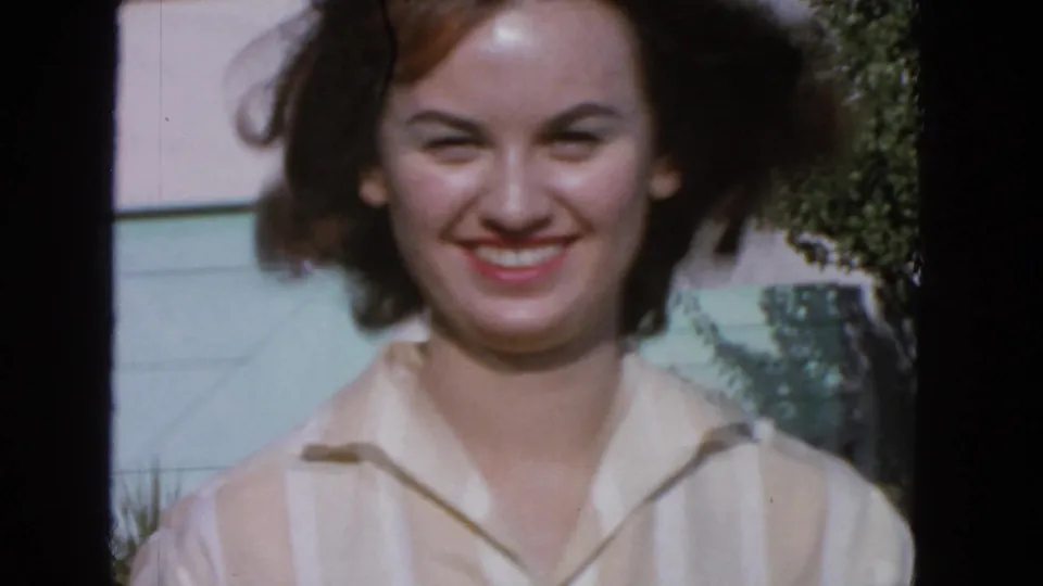 Closer 1963 San Francisco home-movie view of the smiling woman, with brown hair lifted by wind and garden foliage behind her.