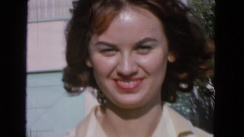 Close-up 8mm frame of the young woman smiling in bright outdoor light, with soft focus and house siding behind her.