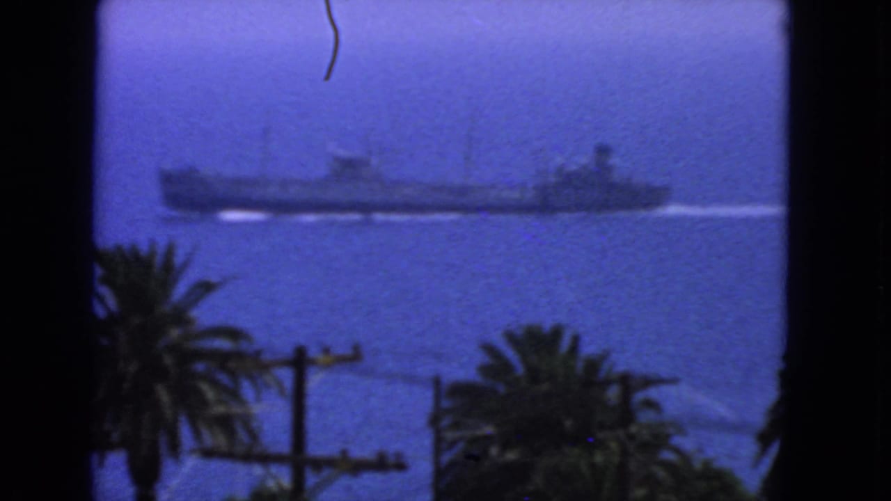 1963 San Francisco 8mm frame showing a large naval vessel on blue coastal water, framed by palms, utility poles, and dark edges.