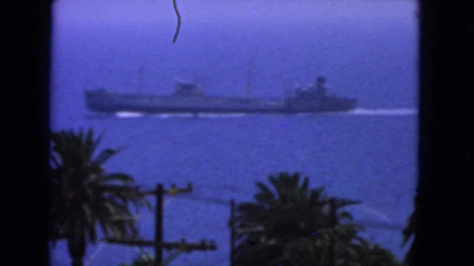 1963 San Francisco 8mm frame showing a large naval vessel on blue coastal water, framed by palms, utility poles, and dark edges.