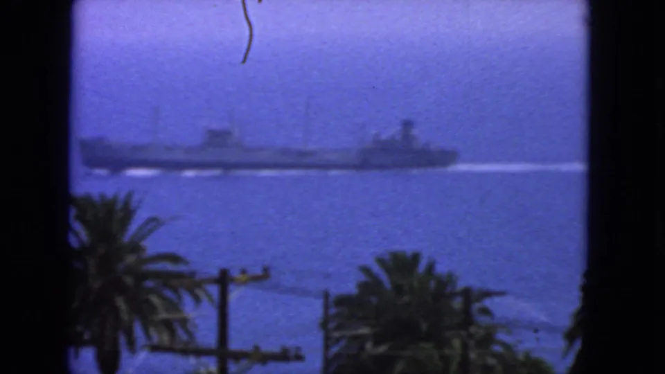 1963 San Francisco home-movie frame of a long naval vessel passing across the bay, partly framed by palm trees and dark window edges.
