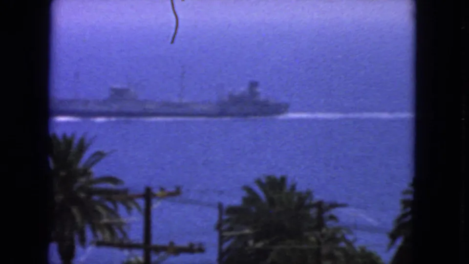 1963 San Francisco 8mm still with the ship partly cropped at left, a white wake on the water, and palms and utility lines below.