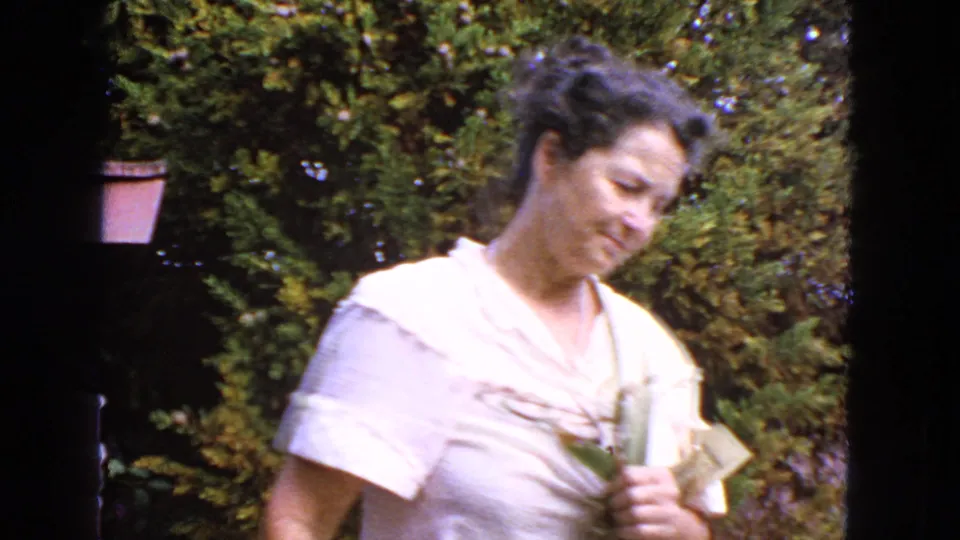 1963 San Francisco home-movie frame showing a woman outdoors in a white blouse holding flowers near garden foliage.