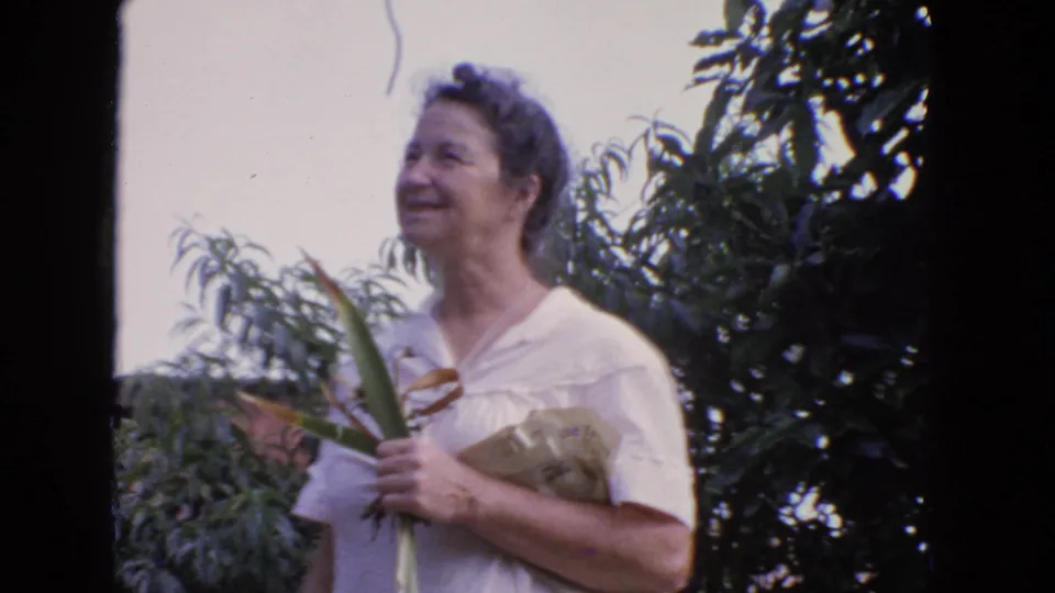 A smiling woman in a light dress holds long leaves and a bundle of greenery in a leafy San Francisco backyard setting.
