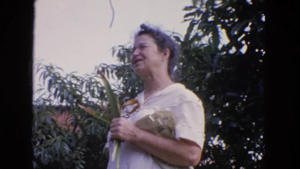 The woman remains in the backyard holding cut plants, framed by soft 8mm focus, foliage, and a low brick wall.
