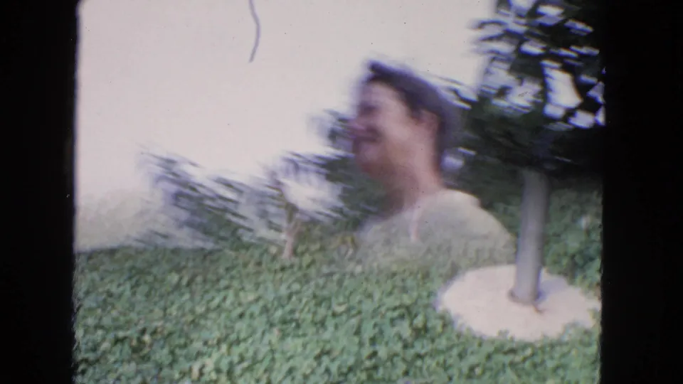 A small dog investigates a pole in the grass while the woman appears motion-blurred in the leafy backyard behind it.
