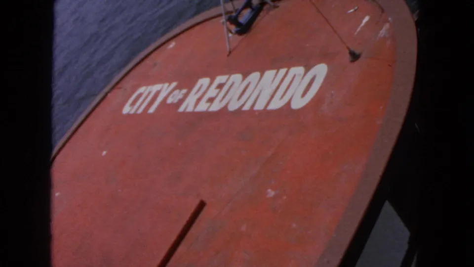 Close angled view of a red circular sign reading "CITY OF REDONDO," with dark film edge, gray backdrop, and visible surface wear.