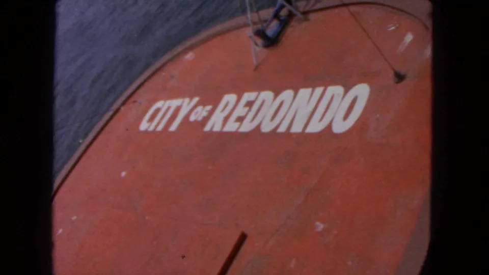 Close 8mm view of the weathered red boat surface marked "CITY OF REDONDO," with dark rippling water visible along the side.