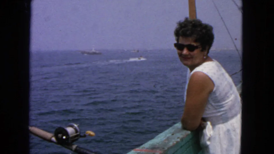 A woman in a white sleeveless dress and dark sunglasses smiles from a boat rail, with boats on the water in the distance.