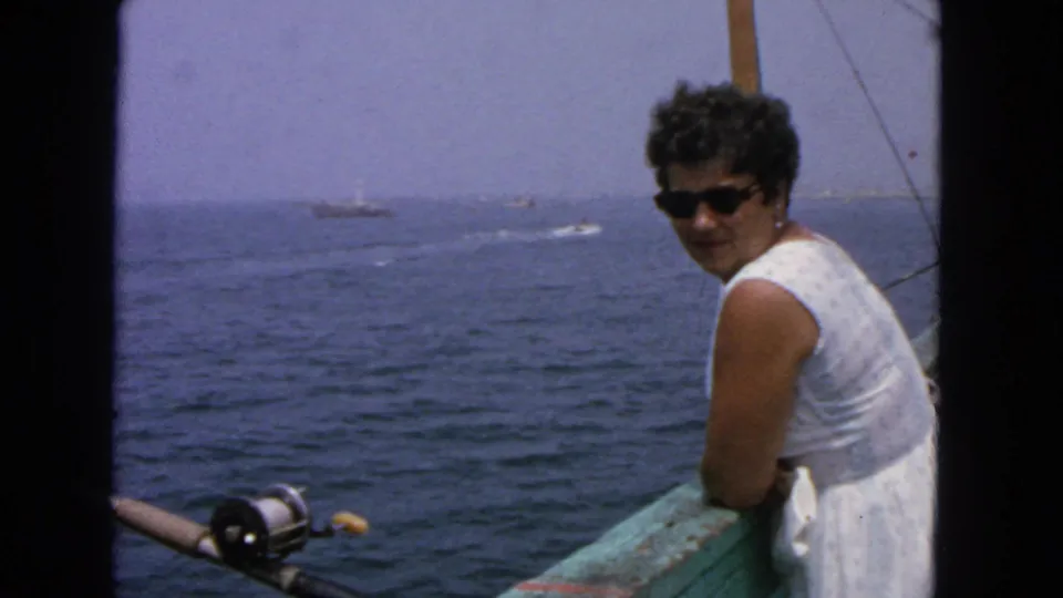The woman looks toward the camera from the boat rail, wearing sunglasses and a white dress against open water and distant boats.