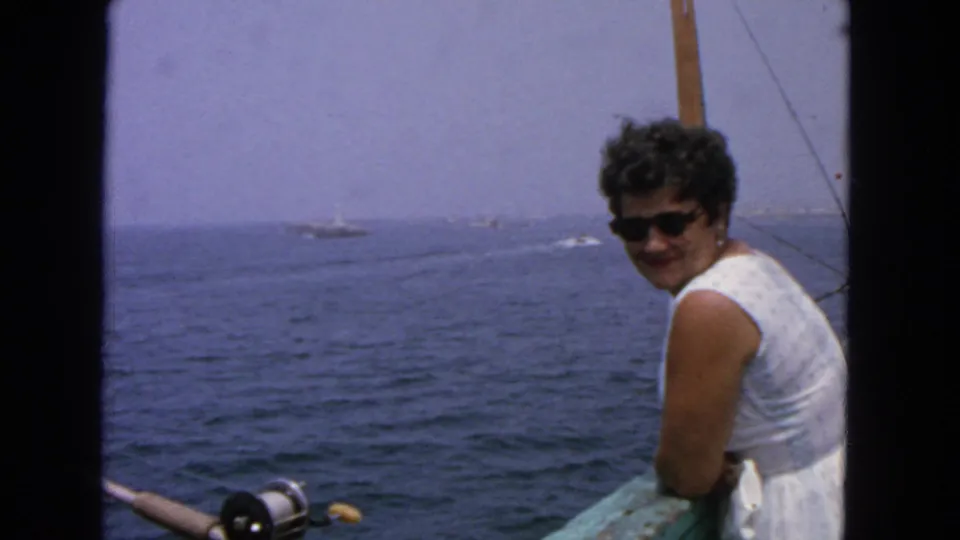 1963 San Francisco-area boating frame with a smiling woman in dark sunglasses, a fishing reel in the foreground, and vessels on the horizon.