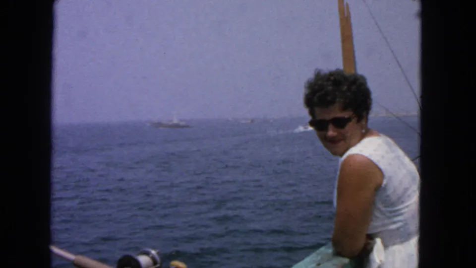 8mm 1963 San Francisco-area home-movie frame of a woman leaning at a boat rail with fishing gear and open water visible.