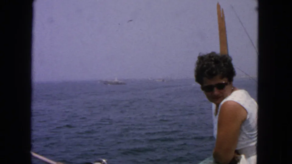 A woman in sunglasses sits near boat rigging while the ocean horizon and small distant vessels appear behind her.