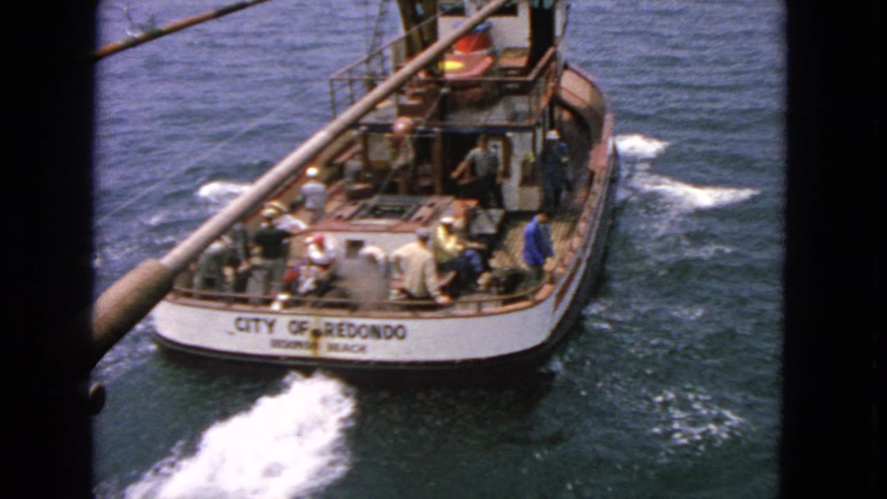 1963 8mm frame of a named fishing boat seen from another vessel off California, with passengers on deck and wake trailing behind.