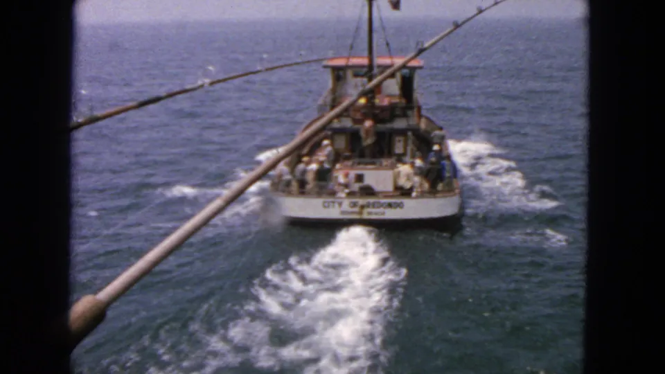 A 1963 8mm view follows a fishing boat on blue water, framed by fishing rods from the filming vessel.