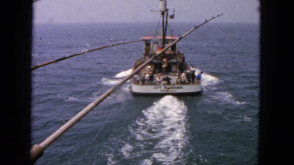 The fishing boat recedes across open water in a 1963 California home-movie frame, with foreground rods emphasizing the filming vessel.