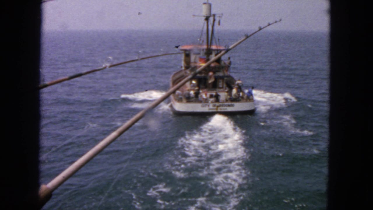 Fishing boat moving across open ocean with rods in the foreground and a white wake behind it.