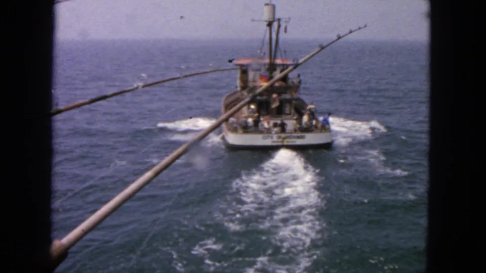 1963 San Francisco-area 8mm frame showing a fishing boat named City of San Francisco moving away across open ocean, seen from another vessel with rods in the foreground.