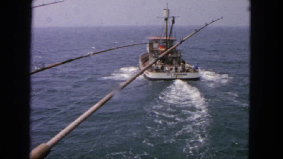 A 1963 8mm view from a second vessel shows the City of San Francisco fishing boat receding over choppy ocean water with fishing rods crossing the foreground.