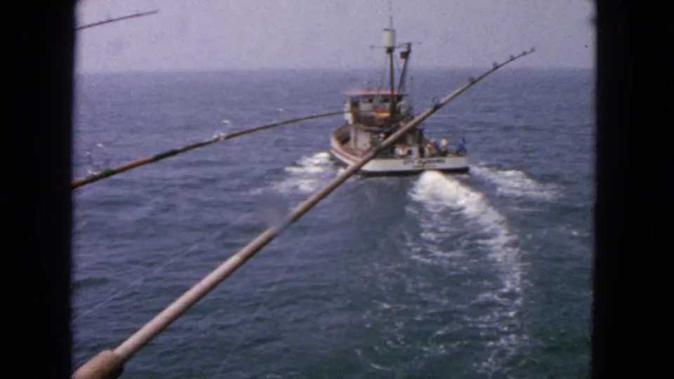 The fishing boat appears farther away in this 1963 San Francisco-area home-movie frame, with long rods in the foreground and a foamy wake marking the path between vessels.