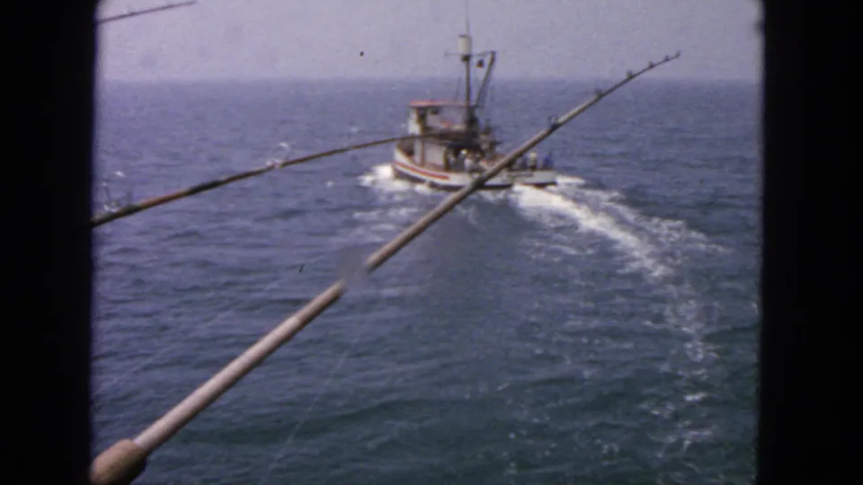 1963 San Francisco home-movie view from a pier as a small boat passes across bay water behind two fishing rods.
