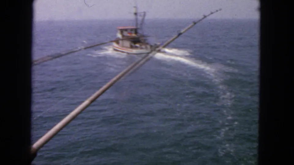 A small boat moves away across San Francisco bay water in this 1963 8mm frame, with fishing rods visible from the pier viewpoint.