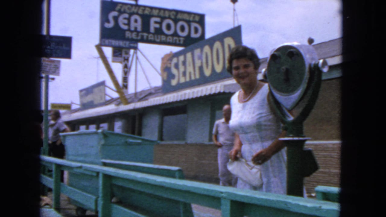 Woman smiles near a coin viewer while a man stands behind her on the pier walkway.