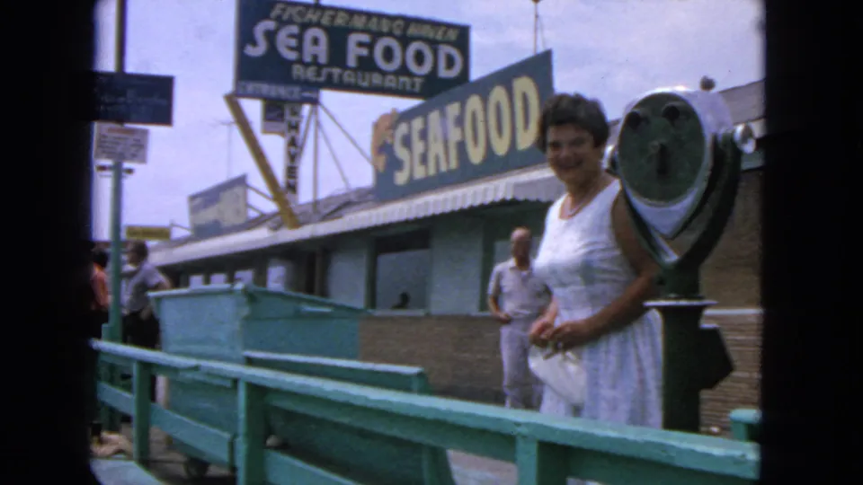 1963 waterfront home-movie frame showing a woman by a pier viewer with seafood restaurant signs above the building.
