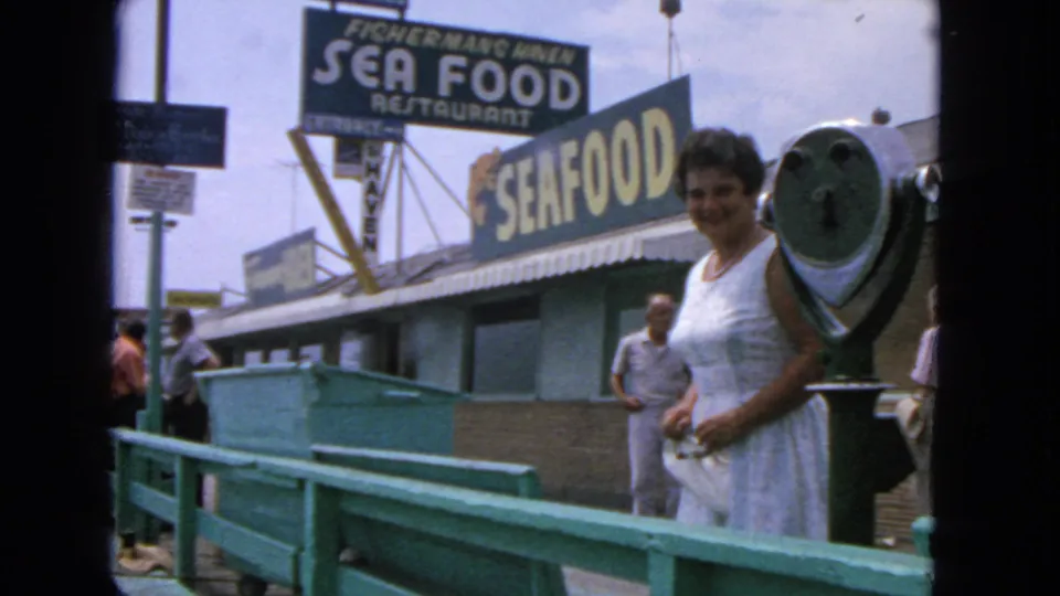 1963 San Francisco pier view with a woman near a green coin viewer, a man behind her, railings, and seafood signs.