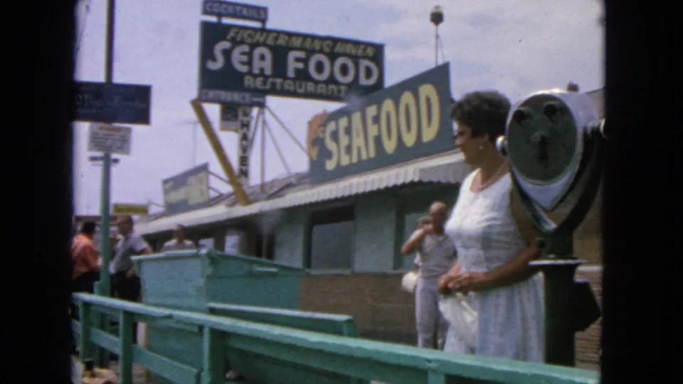 1963 pier scene with a woman beside a coin viewer, turquoise railings, and Fisherman's Haven seafood signage visible.