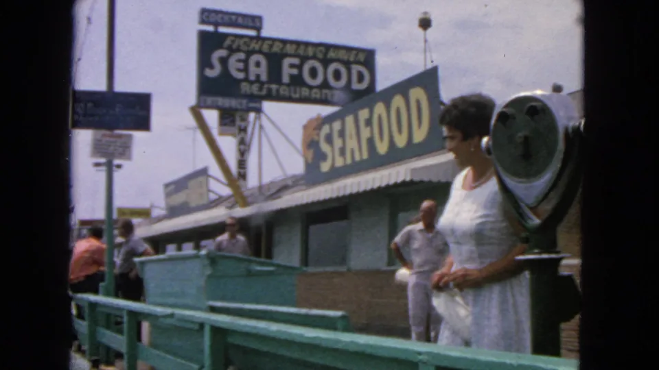 San Francisco 8mm frame of a woman at a waterfront viewer with seafood restaurant signs and casual pier traffic.