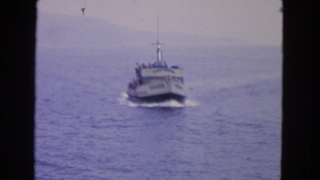 Passenger boat continues toward the camera over open water with shoreline in haze.