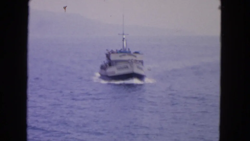 The Voyager moves closer on blue water, with passengers faintly visible on deck and a hazy coastline beyond.