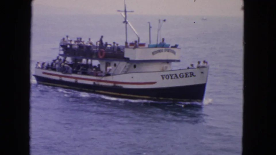 1963 San Francisco home-movie frame showing the white passenger boat Voyager moving across calm water with passengers on deck.