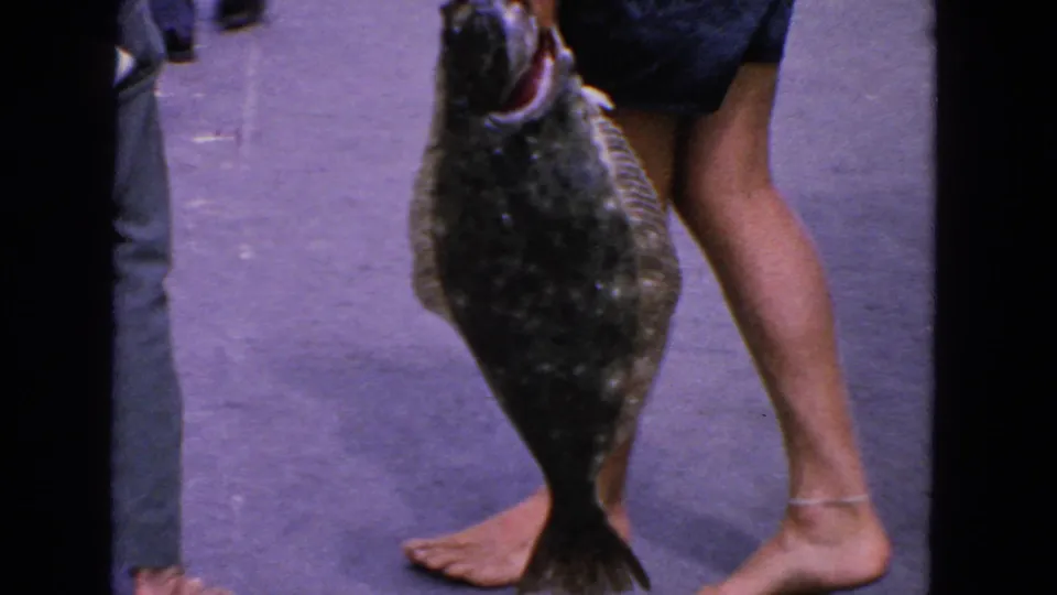 1963 home-movie frame of a person displaying a large flat fish on a waterfront deck or dock.