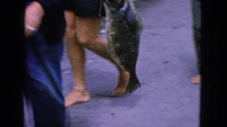 Handheld 1963 San Francisco 8mm frame showing a large flat fish carried beside barefoot dockside onlookers.