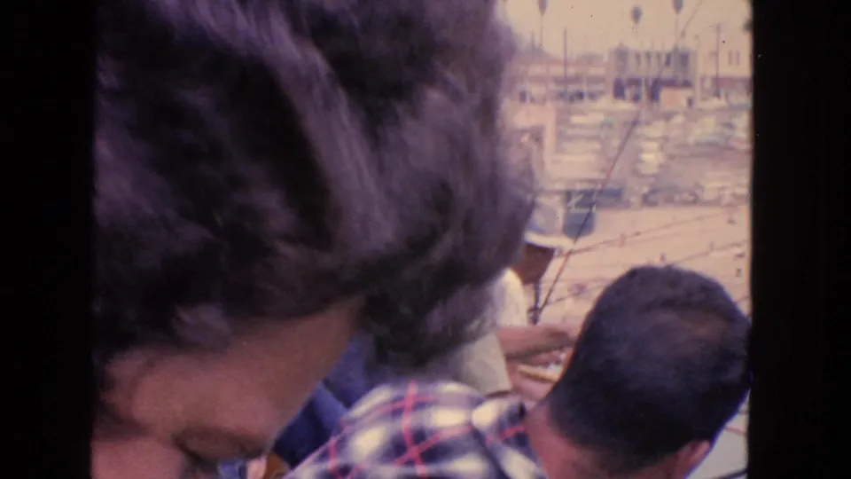 Home-movie view of nearby people at a waterfront, with small boats and marina masts visible in the distance.