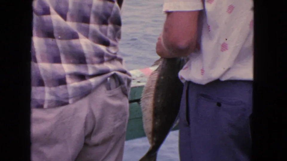 1963 San Francisco home-movie frame showing two men on a pier with a freshly caught fish beside the bay water.