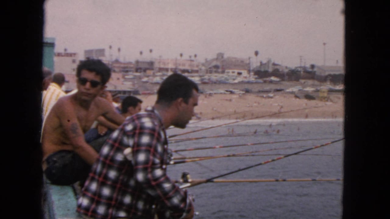 Men sit with fishing rods along a pier above a beach.