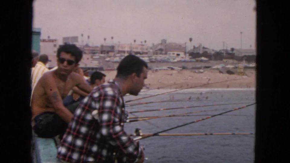 1963 San Francisco waterfront frame with men fishing from a pier, beach and coastal buildings in the distance.