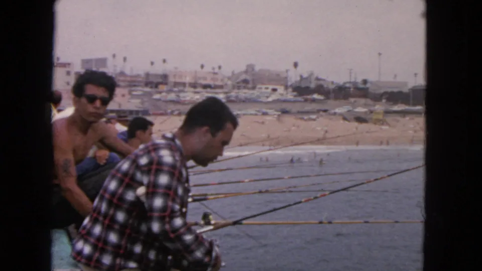 1963 San Francisco home-movie frame of pier anglers, including a man in a red plaid shirt and another shirtless man in sunglasses.