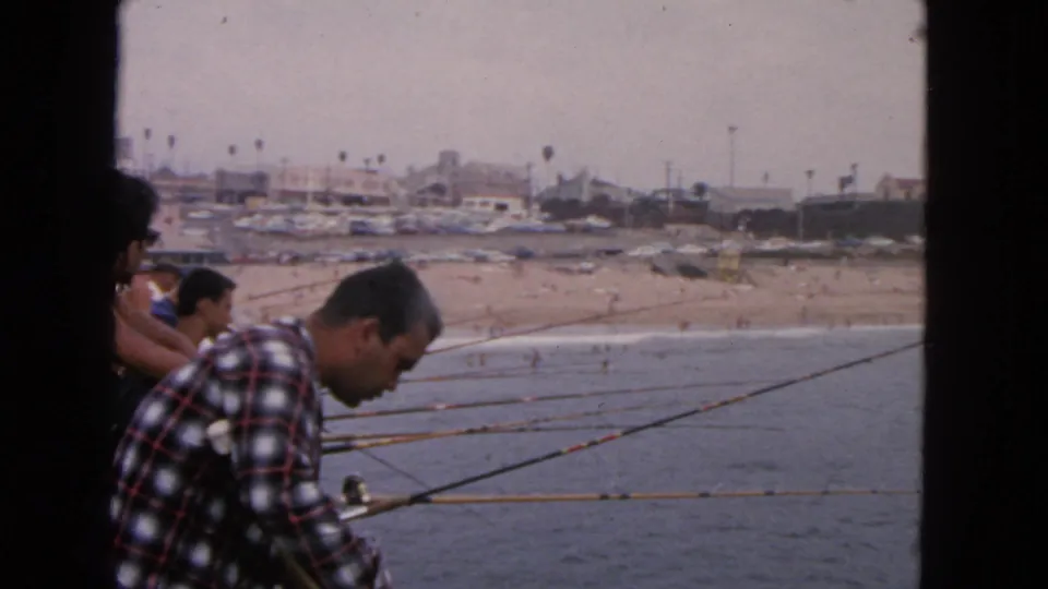 1963 San Francisco waterfront frame showing seated anglers and rods stretching across a pier above the shoreline.