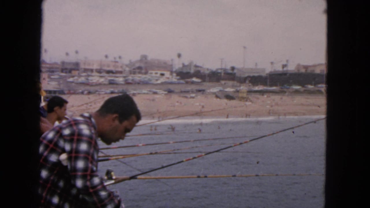 1963 San Francisco 8mm frame showing a man in a plaid shirt fishing from a pier above the ocean, with a crowded beach in the background.