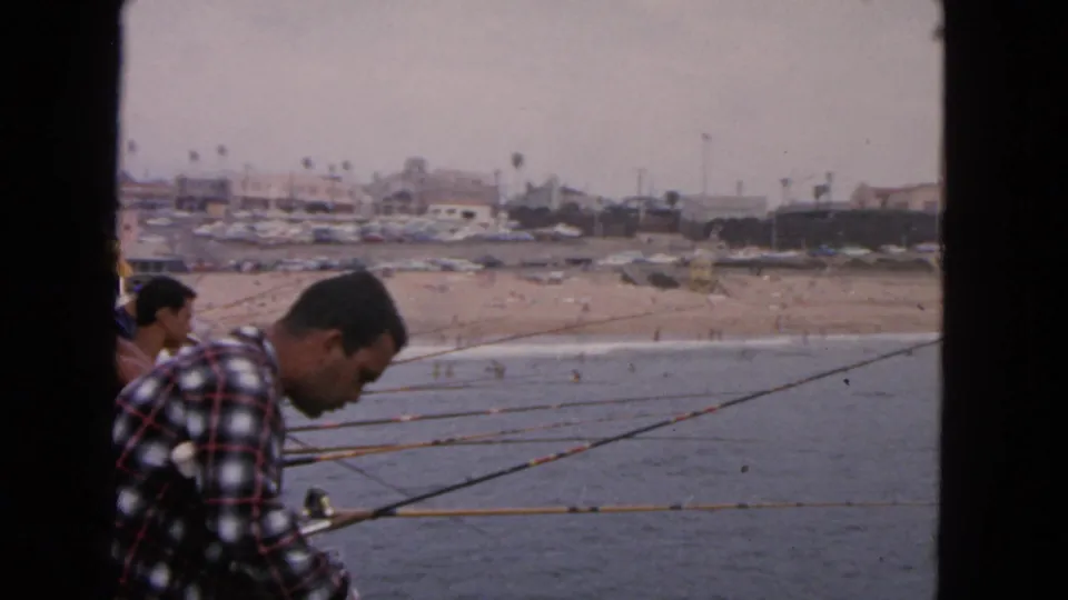 1963 San Francisco 8mm frame showing a man in a plaid shirt fishing from a pier above the ocean, with a crowded beach in the background.