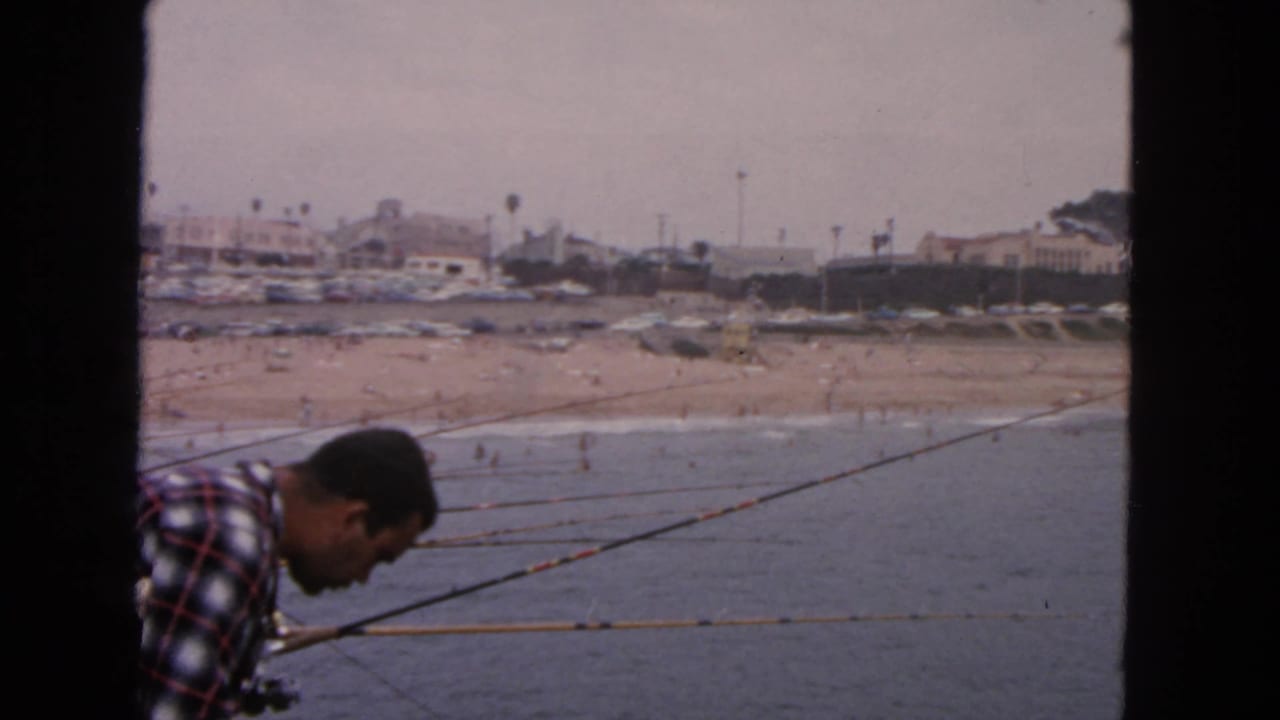 1963 San Francisco home-movie frame of an angler leaning near multiple rods on a pier, with surf, beachgoers, and buildings beyond.