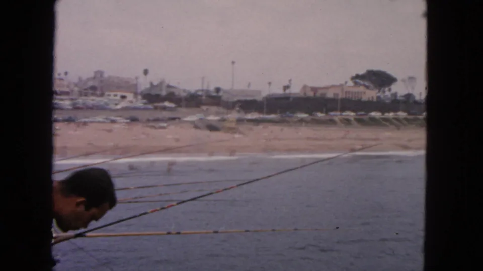 1963 San Francisco 8mm frame with the fisherman partly out of view, rods angled over the water, and a hazy beach and shoreline behind.